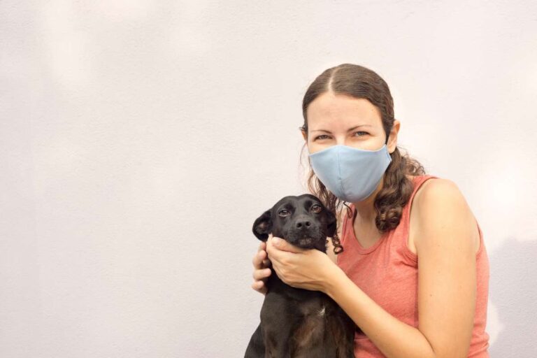 A woman wearing a mask, possibly because she has Covid, and holding a black dog.