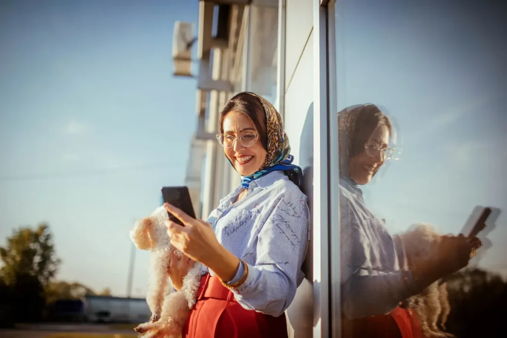 Woman smiling while holding a small dog and using a smartphone outdoors.